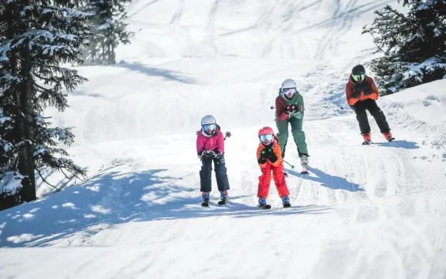 A family skiing downhill together, all schussing, on a groomed piste, with two children leading and adults following, surrounded by snowy trees and bright winter sun.